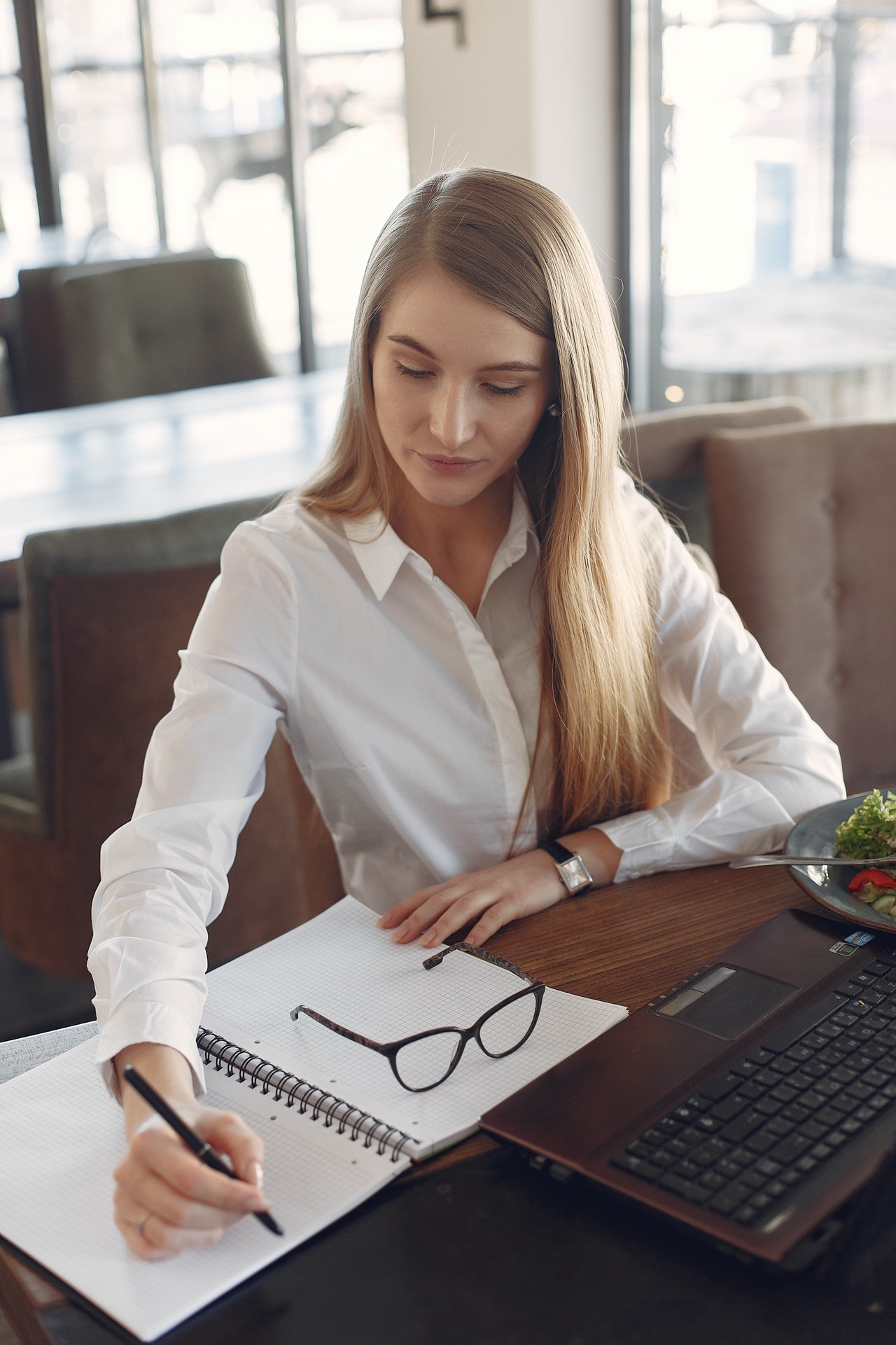 woman seated at large table writing on notepad with laptop in front of her